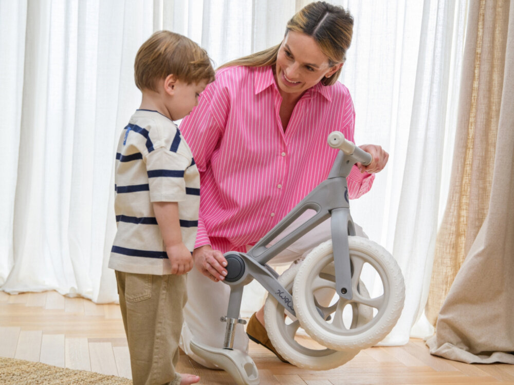 Rowerek biegowy MOMI Noru Szary pokój osoba trzyma w dłoniach złożony rowerek obok stoi dziecko w tle okno składany mechanizm ramy szybkie złożenie przy użyciu jednego przycisku ułatwia przechowywanie prosty system składania