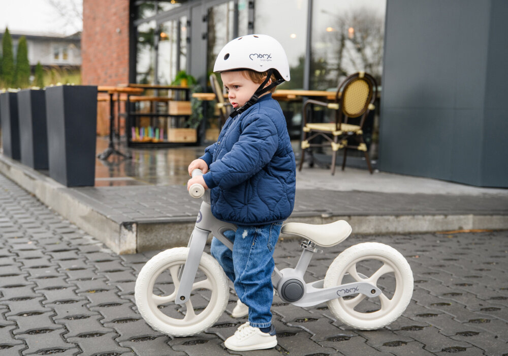 Rowerek biegowy MOMI Noru Szary ulica dziecko stoi przy rowerku w tle taras z ogródkiem przy restauracji szeroka kierownica z gumowymi antypoślizgowymi uchwytami nie ma blokady skrętu kierownica złoży się na płasko minimalizując ryzyko uderzenia ułatwia manewrowanie wpływa na zwrotność rowerka