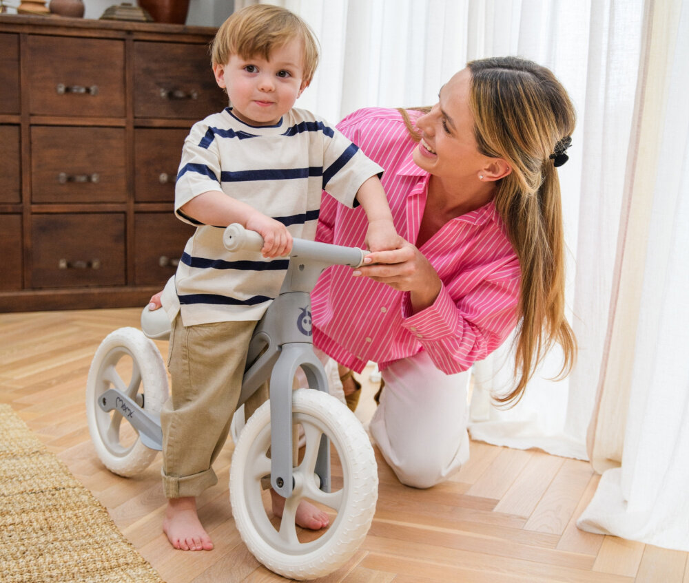 Rowerek biegowy MOMI Noru Szary pokój dziecko siedzi na rowerku obok klęczy dorosła osoba trzyma za rowerek w tle okno komoda spełnia europejskie normy EN 71-1, EN 71-2 i EN 71-3 bezpieczeństwo zastosowanych materiałów i konstrukcji odpowiedni dla dzieci od 2. roku życia do maksymalnej wagi do 25 kg pojazd został sprawdzony pod kątem użytkowania