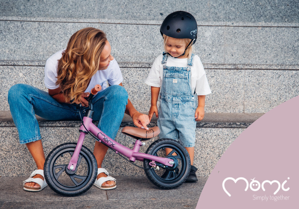Rowerek biegowy MOMI Breki Fioletowy schody betonowe na schodach siedzi dorosła osoba obok stoi dziecko w kasku dorosły trzyma rowerek przed sobą aktywność rozwój pierwszy pojazd dziecka