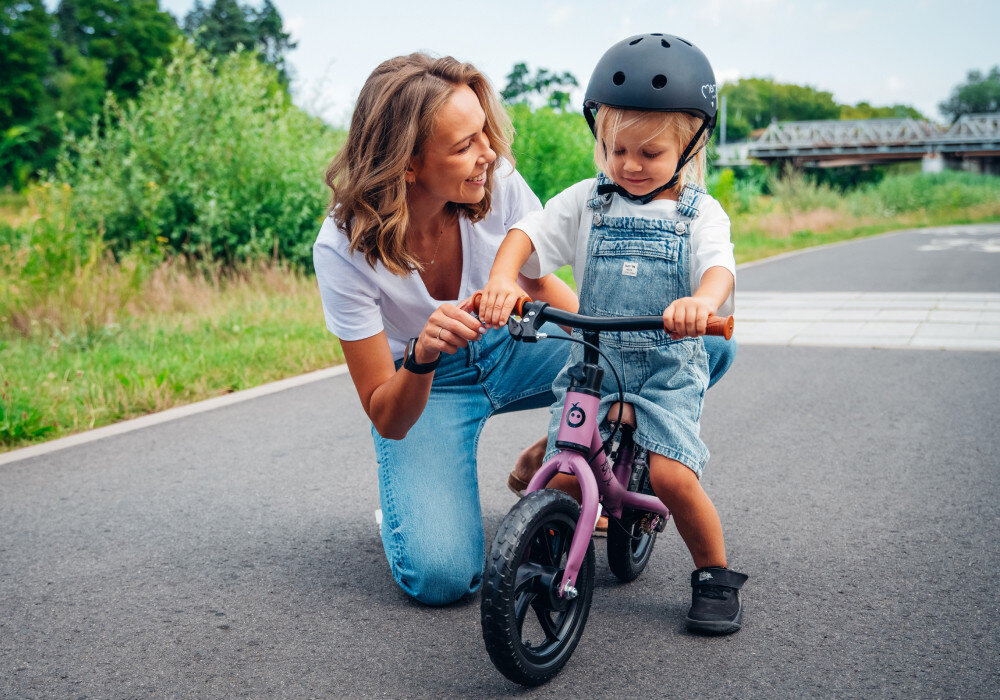 Rowerek biegowy MOMI Breki Fioletowy park droga rowerowa dorosła osoba klęczy obok dziecka stojącego przy rowerku w tle drzewa krzewy niebo most nauka równowagi koordynacji świadomego kontrolowania ruchu przygotowuje malucha do późniejszej jazdy na klasycznym rowerze rozwija motorykę aktywność na świeżym powietrzu lekka konstrukcja