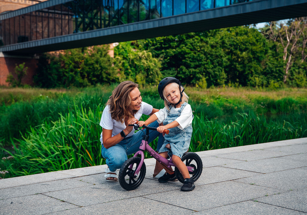 Rowerek biegowy MOMI Breki Fioletowy park droga rowerowa dorosła osoba klęczy obok dziecka stojącego przy rowerku w tle drzewa krzewy niebo most lekka rama o masie 2,25 kg swobodne prowadzenie rowerka bez wysiłku stalowa 8,5-calowa konstrukcja trwałość stabilność ramy wspiera rozwój równowagi