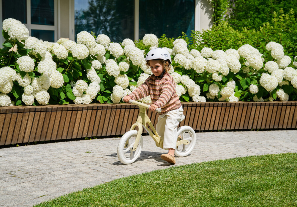 Rowerek biegowy MOMI Noru Żółty park ogród droga dziecko jedzie na rowerku w tle kwiaty okno szeroka kierownica z gumowymi antypoślizgowymi uchwytami nie ma blokady skrętu kierownica złoży się na płasko minimalizując ryzyko uderzenia ułatwia manewrowanie wpływa na zwrotność rowerka