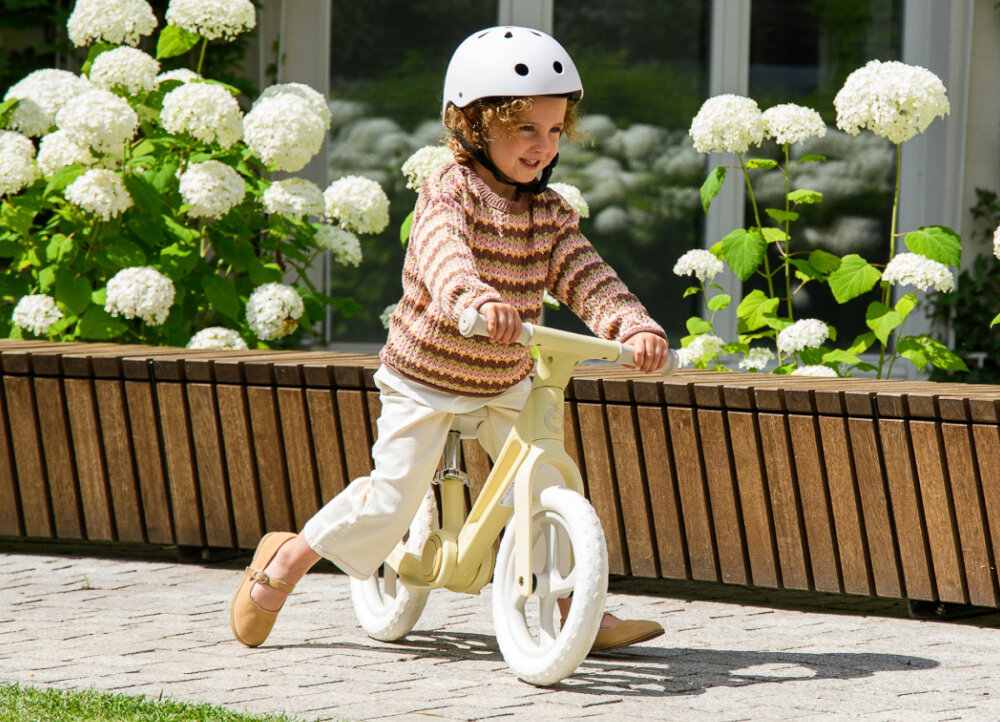 Rowerek biegowy MOMI Noru Żółty park ogród droga dziecko jedzie na rowerku w tle kwiaty okno spełnia europejskie normy EN 71-1, EN 71-2 i EN 71-3 bezpieczeństwo zastosowanych materiałów i konstrukcji odpowiedni dla dzieci od 2. roku życia do maksymalnej wagi do 25 kg pojazd został sprawdzony pod kątem użytkowania