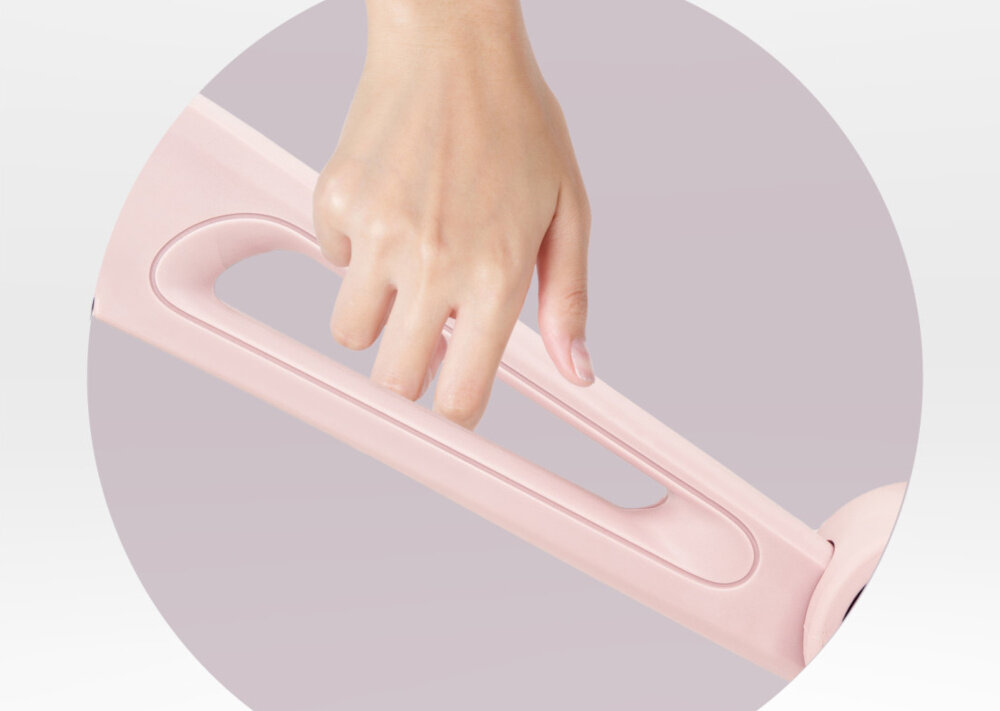 Rowerek biegowy MOMI Noru Różowy zbliżenie na ramę osoba trzyma dłoń na otworze 10-calowa rama z wytrzymałego nylonu trwałość przy zachowaniu niskiej wagi 3 kg wygodny otwór łatwe przenoszenie rowerka ułatwia transport i przechowywanie