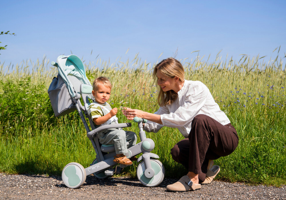 Rowerek trójkołowy MOMI Solivia 4w1 Opony Eva Zielony rączka droga łąka dziecko siedzi na rowerku obok klęczy dorosła osoba w tle pole dla dzieci od 12. miesiąca życia nauka równowagi koordynacji samodzielnej jazdy zachęca do ruchu i aktywności na świeżym powietrzu