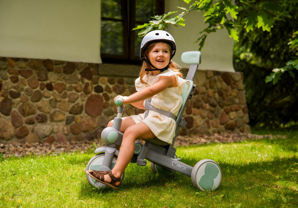 Rowerek trójkołowy MOMI Solivia 4w1 Opony Eva Zielony rączka trawnik dziecko siedzi na rowerku zapięte pasami w tle dom drzewa pięciopunktowe pasy z możliwością demontażu pałąk funkcja wolnej kierownicy kontrola nad jazdą regulowany daszek chroni przed słońcem możliwość jego całkowitego zdjęcia komfort dziecka