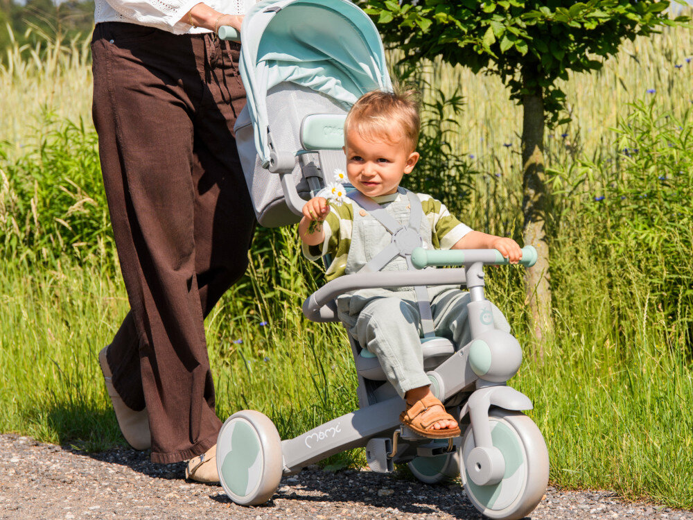Rowerek trójkołowy MOMI Solivia 4w1 Opony Eva Zielony rączka droga dziecko siedzi na rowerku dorosła osoba pcha rowerek przed sobą w tle rośliny drzewa teleskopowy uchwyt dla rodzica trzystopniowa regulacja w zakresie 87/92/97 cm funkcja wolnego koła rodzicowi pełna kontrola nad kierunkiem jazdy