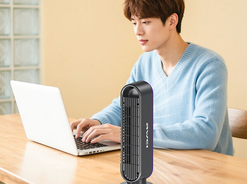 Osoba siedzi przy drewnianym stole, pracując na laptopie. Obok znajduje się szary wentylator z widocznym logo \'AWEI\'.