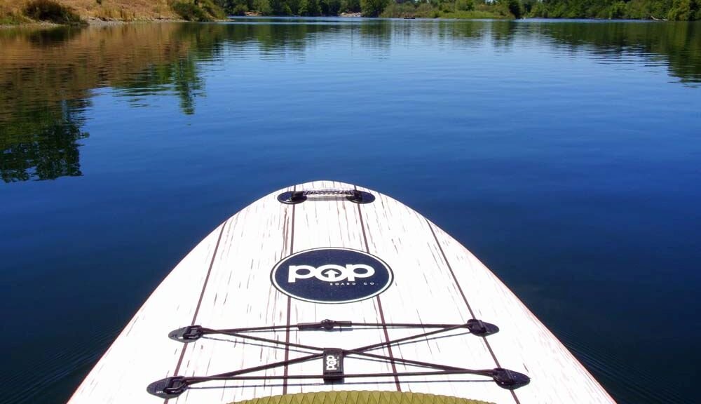 Deska do paddleboardingu na spokojnej wodzie z widocznym logo \'POP\' na powierzchni. W tle widać zalesiony brzeg i odbicie drzew w wodzie.