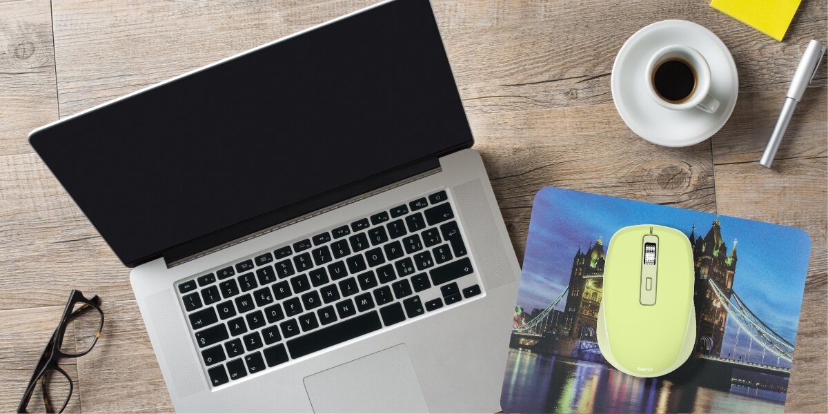 Limonkowa mysz na podkładce z motywem mostu, leżąca na drewnianym biurku obok laptopa, kawy i okularów.