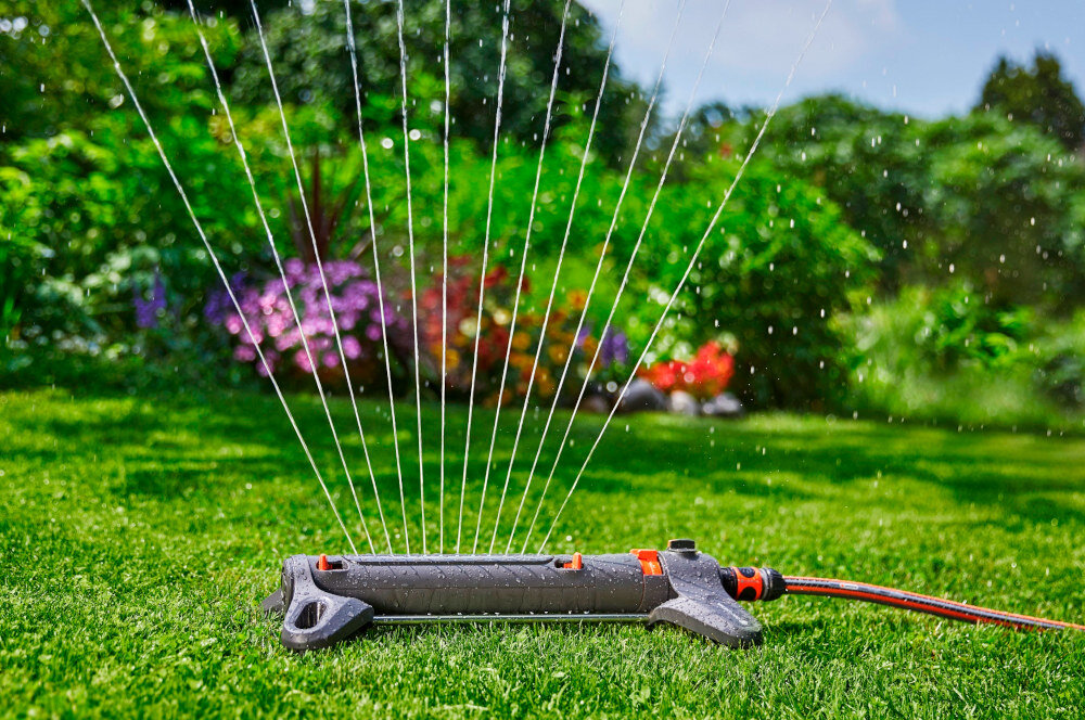 Zraszacz GARDENA Aquazoom S 18710-20 trawnik zraszacz na trawniku w tle drzewa krzewy kwiaty zraszacz wahadłowy do równomiernego nawadniania trawników i ogrodów o powierzchni od 9 do 150 m² eliminuje potrzebę ręcznego podlewania oszczędność czasu i energii