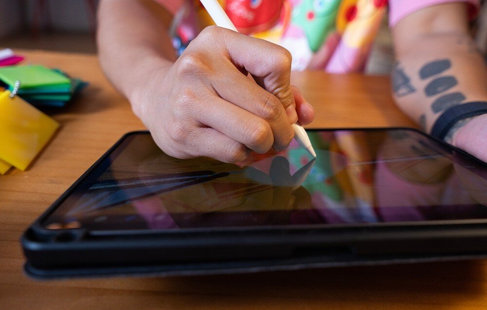 Osoba rysuje na tablecie za pomocą rysika. W tle widoczne są kolorowe elementy i fragmenty ubrania.