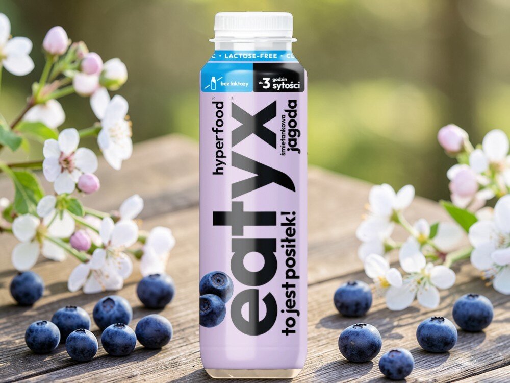 Posiłek w płynie EATYX Hyperfood Śmietankowo-jagodowy (300 ml) jasne tło butelka ustawiona na drewnianym blacie wokół butelki owoce jagód i białe kwiatki posiłek w płynnie zbilansowany posiłek zdrowie