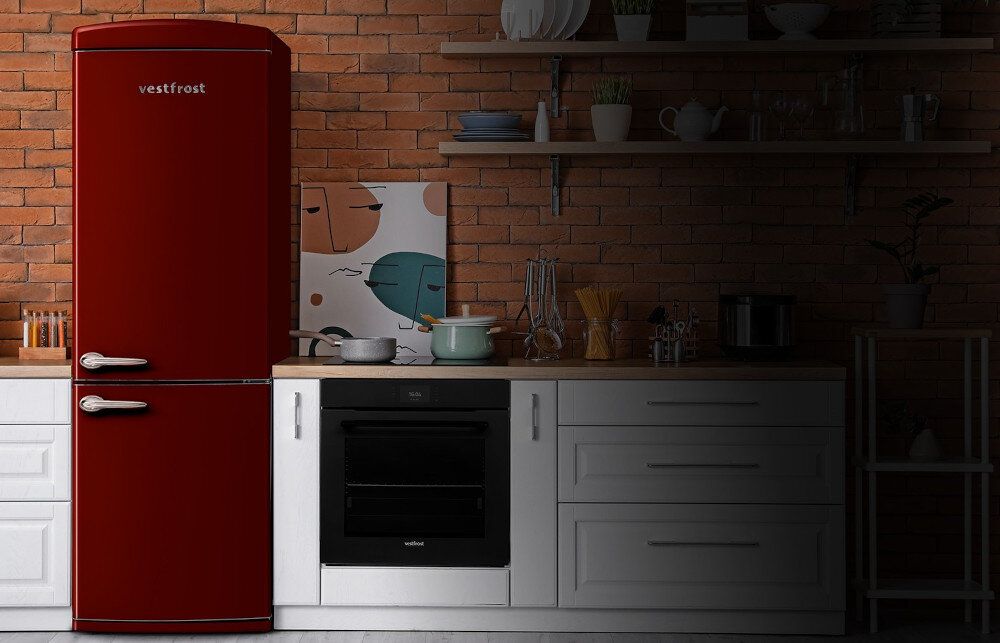 Lodówka VESTFROST VR-FB373-2E2BR Zdjęcie pokazuje bordową lodówko‑zamrażarkę Vestfrost w stylu retro ustawioną w jasnej kuchni z białą zabudową i ceglaną ścianą w tle. Oświetlenie skupia się na lodówce i blacie z garnkami oraz akcesoriami kuchennymi, podczas gdy reszta wnętrza pozostaje w półmroku, podkreślając kolor i design urządzenia