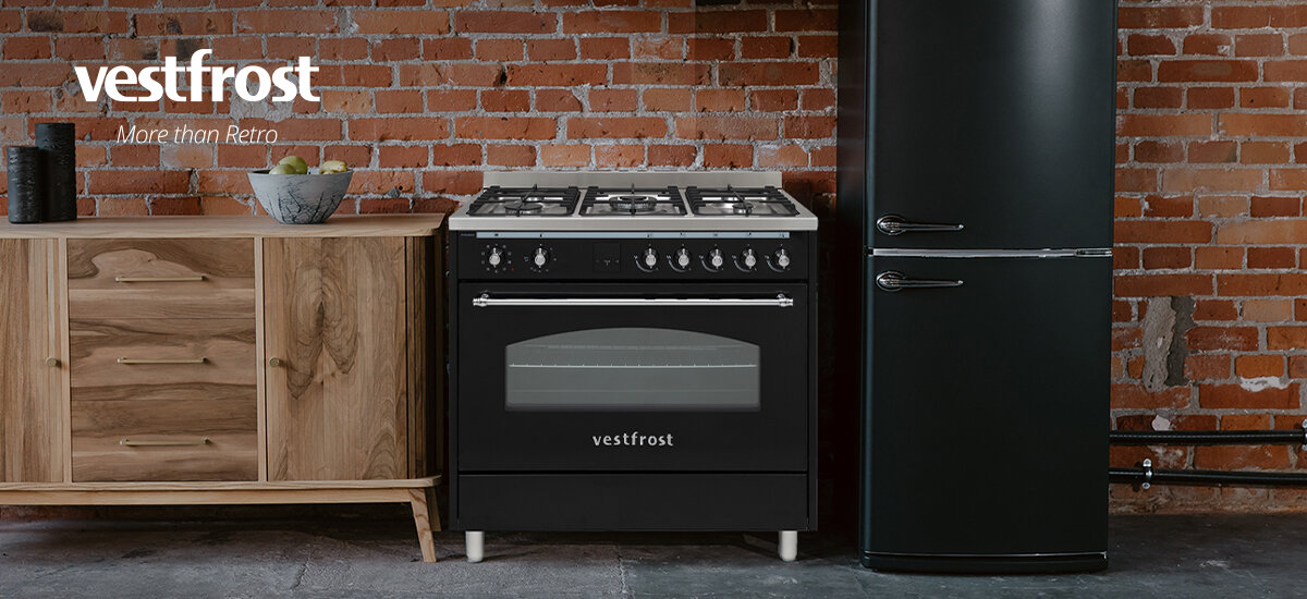 Kuchnia VESTFROST VC-993WNPBK.2 Zdjęcie przedstawia czarną kuchenkę gazową na tle ceglanej ściany, ustawioną obok stylowej lodówki retro. Całość utrzymana jest w industrialnym, loftowym stylu z drewnianą komodą po lewej stronie. Połączenie czerni i naturalnego drewna nadaje wnętrzu elegancji oraz klasycznego charakteru.
