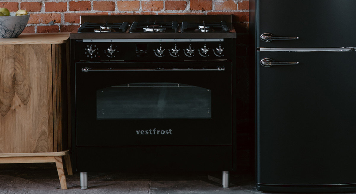 Kuchnia VESTFROST VC-993WNPBK.2 Na zdjęciu widać kuchenkę, lodówkę i drewnianą komodę, ukazane w szerszym ujęciu z logotypem marki i hasłem w górnym rogu. Wnętrze charakteryzuje się ceglaną ścianą, wyeksponowanymi rurami i loftowym klimatem. Kompozycja podkreśla unikalny styl retro połączony z funkcjonalnością sprzętów design czarno satynowe wykończenie styl retro elegancki wygląd technologie klasyczny styl praktyczność estetyka Vestfrost retro