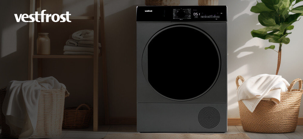 Suszarka VESTFROST VT-FD4EHY70EU Na zdjęciu widoczna jest suszarka bębnowa Vestfrost o ciemnym froncie, ustawiona w stylowym, jasnym pomieszczeniu. Po bokach stoją wiklinowe kosze z ręcznikami, a wnętrze jest uporządkowane i dobrze oświetlone naturalnym światłem