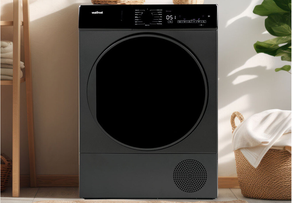 Suszarka VESTFROST VT-FD4EHY70EU Na zdjęciu znajduje się suszarka bębnowa Vestfrost w ciemnym kolorze, umieszczona w jasnym, eleganckim wnętrzu. Urządzenie stoi pomiędzy półką z ręcznikami a koszem, a obok widoczna jest roślina doniczkowa, co podkreśla przytulny charakter aranżacji suszarka do domu elegancka suszarka z wyświetlaczem suszarka premium