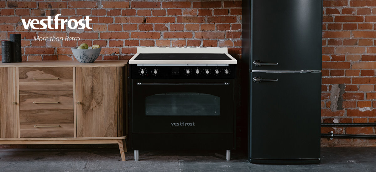 Kuchnia VESTFROST VC-993WNPIBK.2 Zdjęcie przedstawia czarną kuchenkę gazową na tle ceglanej ściany, ustawioną obok stylowej lodówki retro. Całość utrzymana jest w industrialnym, loftowym stylu z drewnianą komodą po lewej stronie. Połączenie czerni i naturalnego drewna nadaje wnętrzu elegancji oraz klasycznego charakteru.
