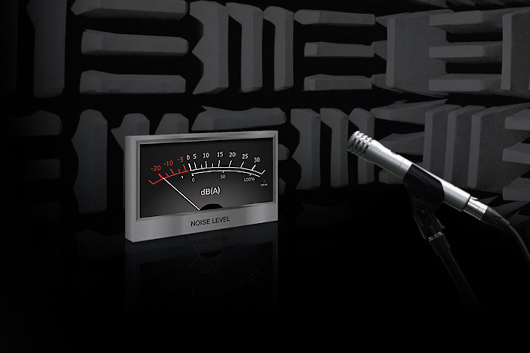Na obrazie widać miernik poziomu hałasu z oznaczeniami w decybelach (dB(A)) oraz mikrofon. Tło stanowi ściana z materiałem tłumiącym dźwięk.