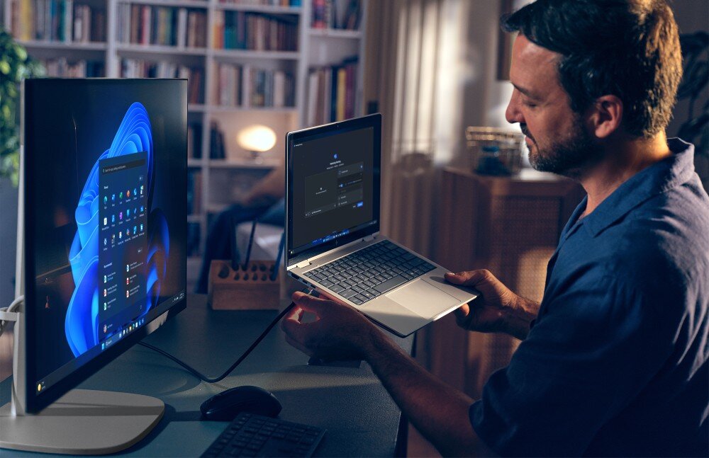 Komputer All in One HP EliteStudio 8 G1I SL Mężczyzna siedzący przy biurku w wieczornym oświetleniu podłącza kablem srebrny laptop do monitora typu All in One, Thunderbolt, Device Switch