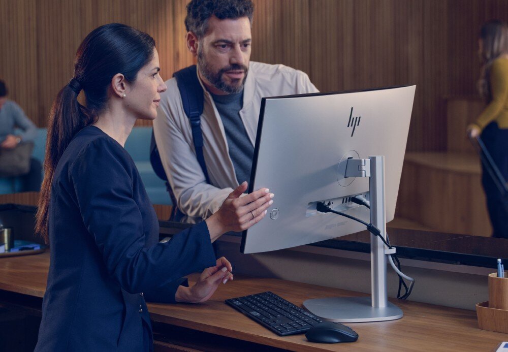 Komputer All in One HP EliteStudio 8 G1I SL Kobieta w ciemnym stroju i mężczyzna w beżowej kurtce wspólnie patrzą na srebrny monitor ustawiony na drewnianym blacie recepcji, łączność bezprzewodowa, Wi-Fi, Bluetooth
