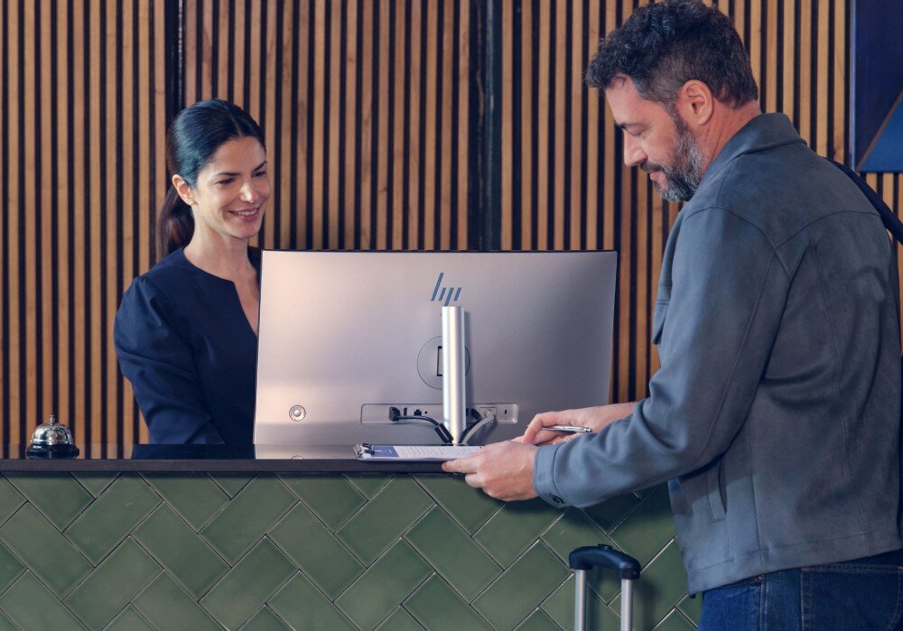Komputer All in One HP EliteStudio 8 G1I SL Tył srebrnego monitora na biurku recepcji, przy którym kobieta w ciemnym stroju obsługuje mężczyznę podpisującego dokumenty, cechy, przeznaczenie, system