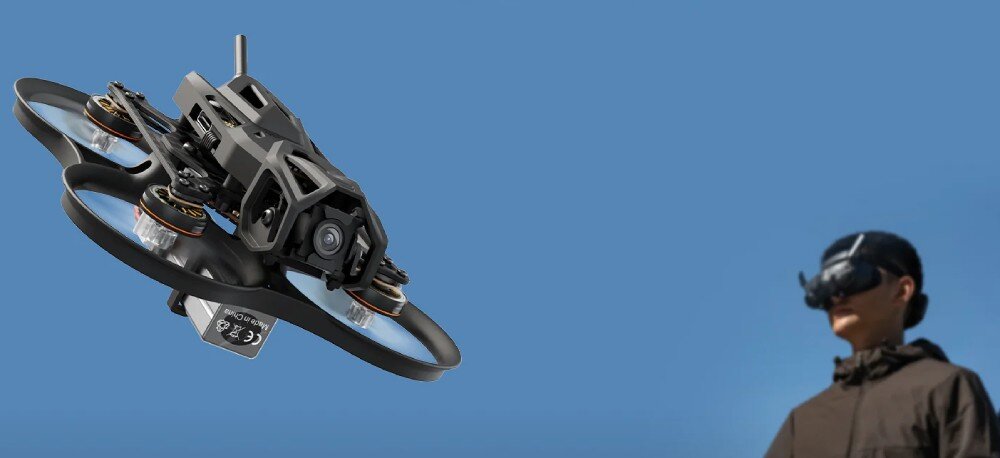 Dron BETAFPV Pavo Femto Dron unoszący się w powietrzu na tle błękitnego nieba i niewyraźnej sylwetki pilota w goglach