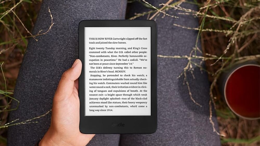 Osoba trzyma czytnik e-booków z widocznym tekstem na ekranie. W tle widać trawę i kubek z napojem.