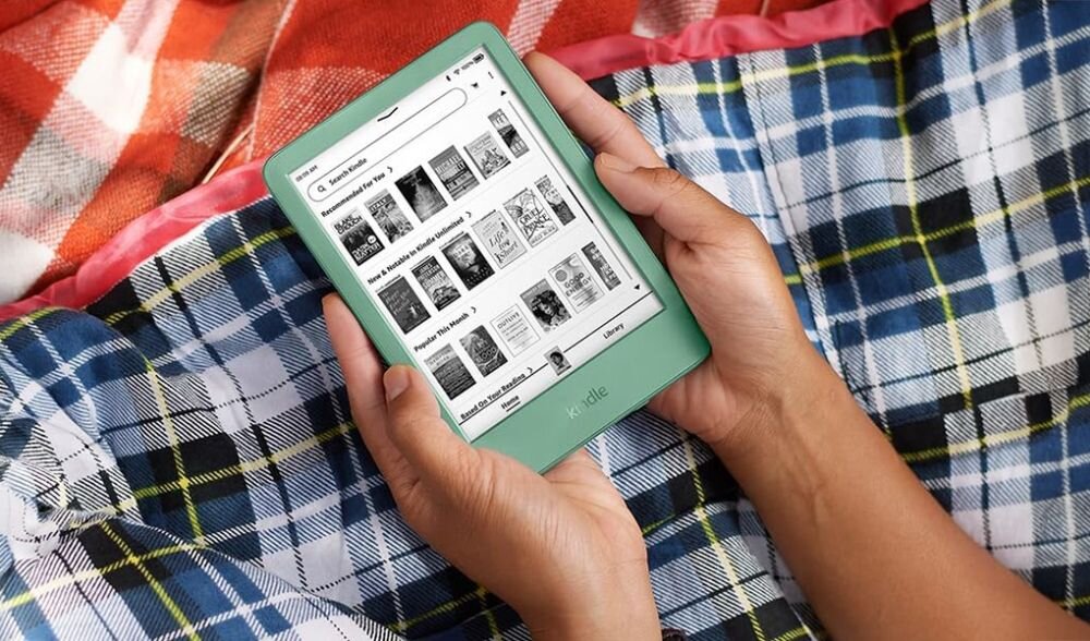 Osoba trzyma zielony czytnik e-booków z widocznym ekranem wyświetlającym okładki książek. W tle znajduje się kolorowy koc w kratę.