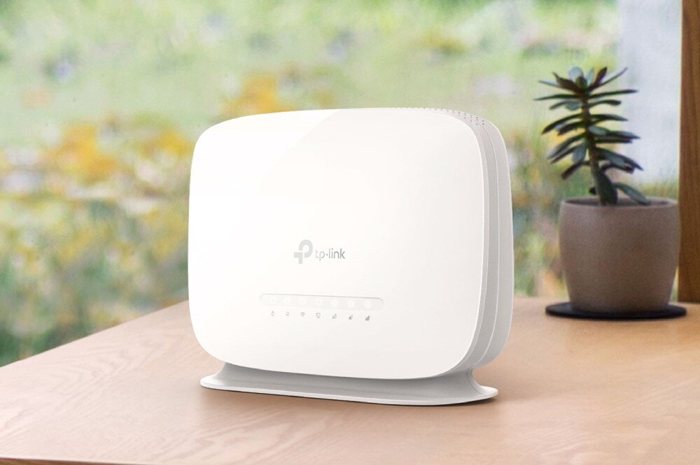Na drewnianym stole stoi biały router z logo \'tp-link\' na przedniej części. W tle widać rozmytą roślinę w doniczce i okno z widokiem na zewnątrz.