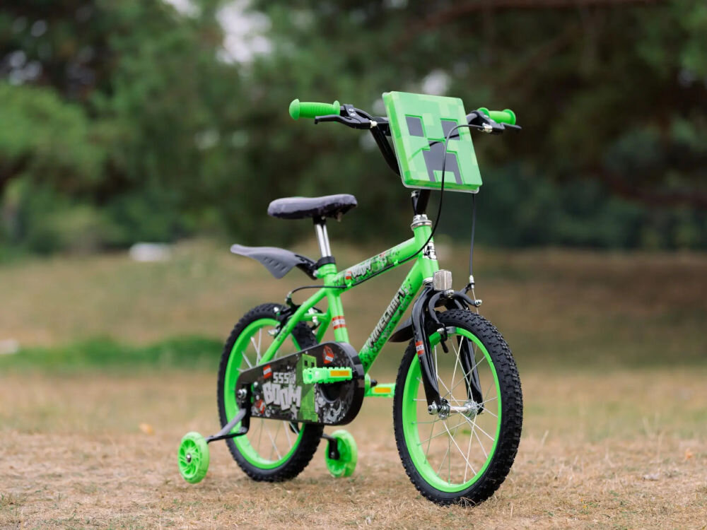 Zielony rower dziecięcy z motywem Minecraft, wyposażony w boczne kółka i tabliczkę z przodu. Rower stoi na trawie, w tle widać drzewa.