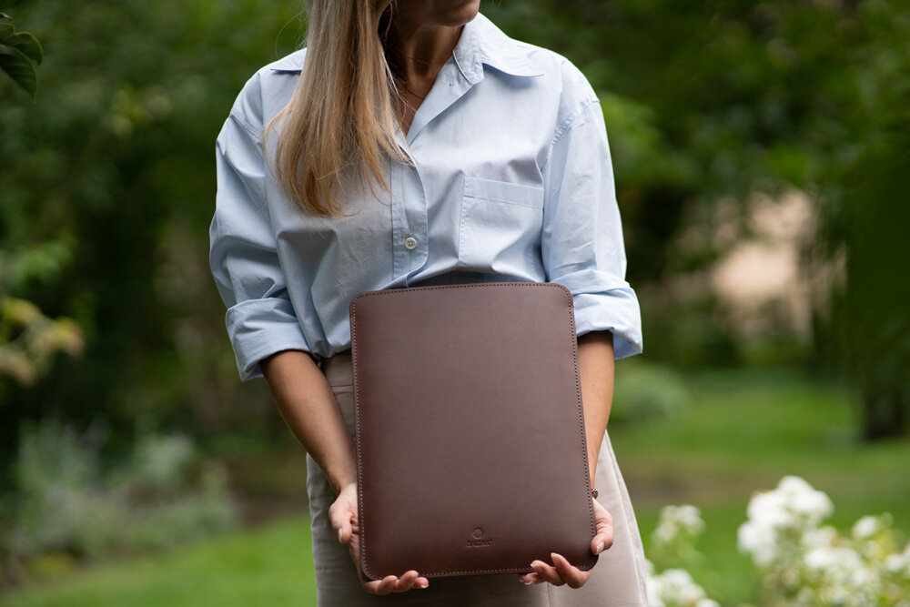 Etui na laptopa BALTAN Sleeve do Apple MacBook Air M4 15.3 cali Drewniany stolik ogrodowy z brązowym skórzanym etui na laptopa oraz laptopem z widocznym logo Apple; obok szklanka z różowymi i czerwonymi kwiatami oraz trzy ceramiczne figurki ptaków.
