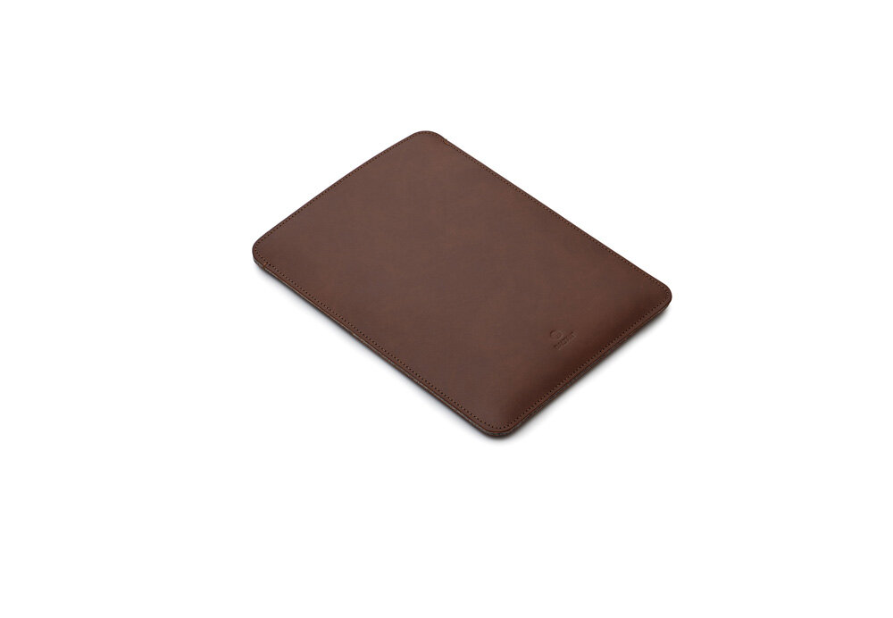 Etui na laptopa BALTAN Sleeve do Apple MacBook Air M4 15.3 cali Jasnobrązowy łatwość czyszczenia prostota pielęgnacja Kobieta w jasnoniebieskiej koszuli i jasnej spódnicy stoi w ogrodzie, trzyma brązowe skórzane etui na laptopa, widoczny fragment sylwetki od ramion w dół, w tle zieleń i białe kwiaty.