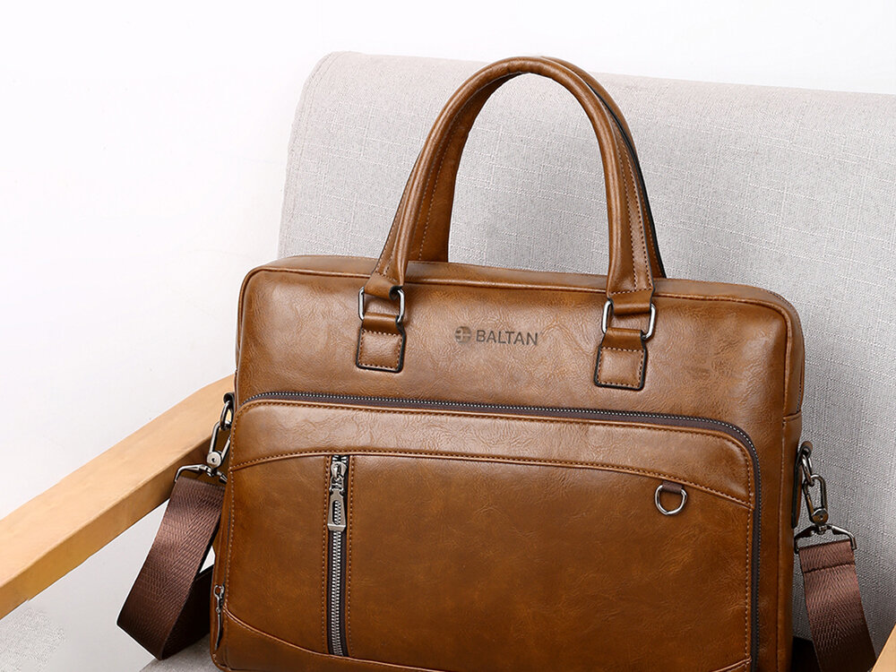 Torba na laptopa BALTAN Office 15.6 cali Jasnobrązowy Brązowa torba na laptopa marki Baltan stojąca na jasnym, tapicerowanym fotelu; widoczne dwa uchwyty, pasek naramienny, przednia kieszeń z zamkiem i metalowy element.