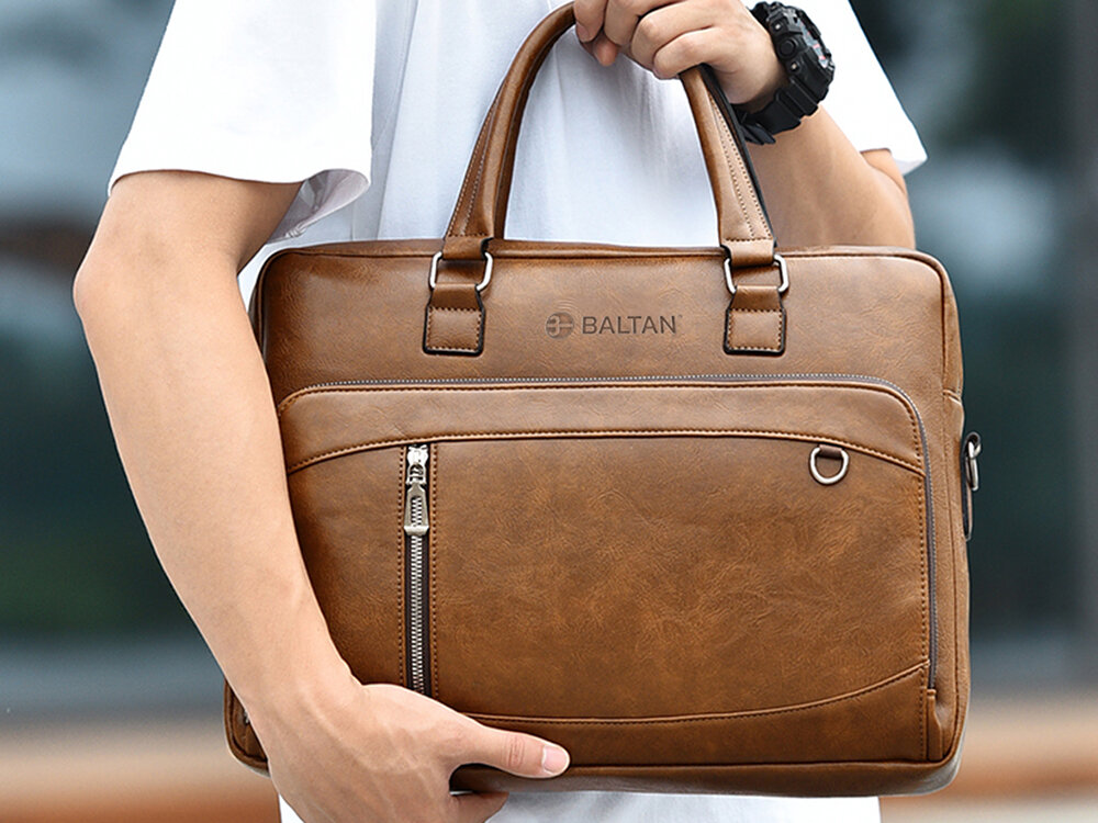 Torba na laptopa BALTAN Office 15.6 cali Mężczyzna w jasnej koszulce i szarych spodniach niesie czarną torbę na laptopa marki Baltan na pasku naramiennym; widoczna przednia kieszeń z zamkiem i metalowym elementem