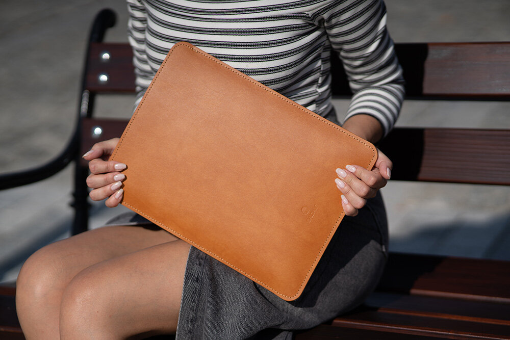 Etui na laptopa BALTAN Sleeve do Apple MacBook Air M4 13.6 cali Jasnobrązowy Razor dla MacBook Air M4 eleganckie design Kobieta siedząca na drewnianej ławce, ubrana w bluzkę w biało-czarne paski oraz ciemną spódnicę, trzyma w dłoniach jasnobrązowe skórzane etui na laptopa. Widać fragment sylwetki od ramion w dół.