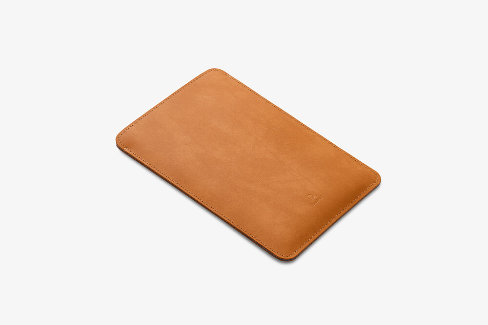Etui na laptopa BALTAN Sleeve do Apple MacBook Air M4 13.6 cali Jasnobrązowy łatwość czyszczenia prostota pielęgnacja Kobieta siedząca na ławce na zewnątrz, ubrana w bluzkę w biało-czarne paski i ciemną spódnicę, wsuwająca laptopa do jasnobrązowego skórzanego etui. Widoczny fragment sylwetki od torsu w dół oraz nogi w delikatnych sandałkach.
