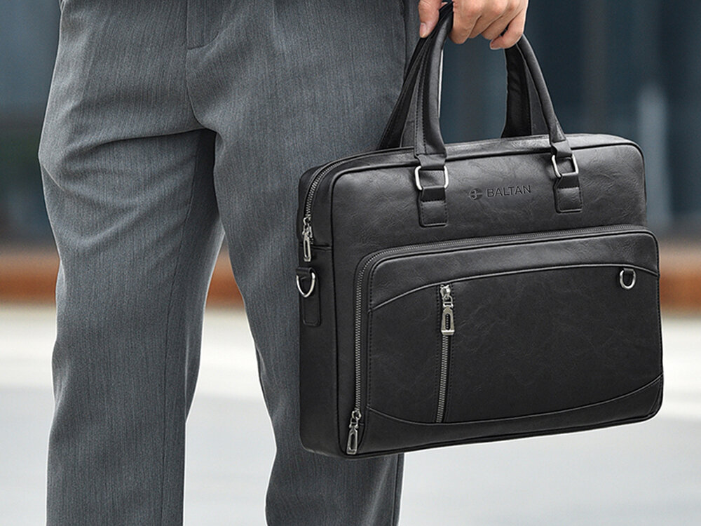 Torba na laptopa BALTAN Office 15.6 cali Mężczyzna w szarych spodniach niesie czarną torbę na laptopa marki Baltan trzymając za uchwyty; widoczny cały przód torby oraz metalowe elementy