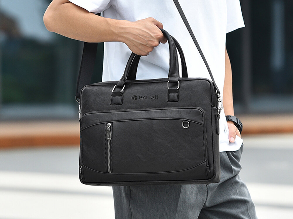 Torba na laptopa BALTAN Office 15.6 cali Mężczyzna w jasnej koszulce i szarych spodniach niesie czarną torbę na laptopa marki Baltan na pasku naramiennym; widoczna przednia kieszeń z zamkiem i metalowym elementem