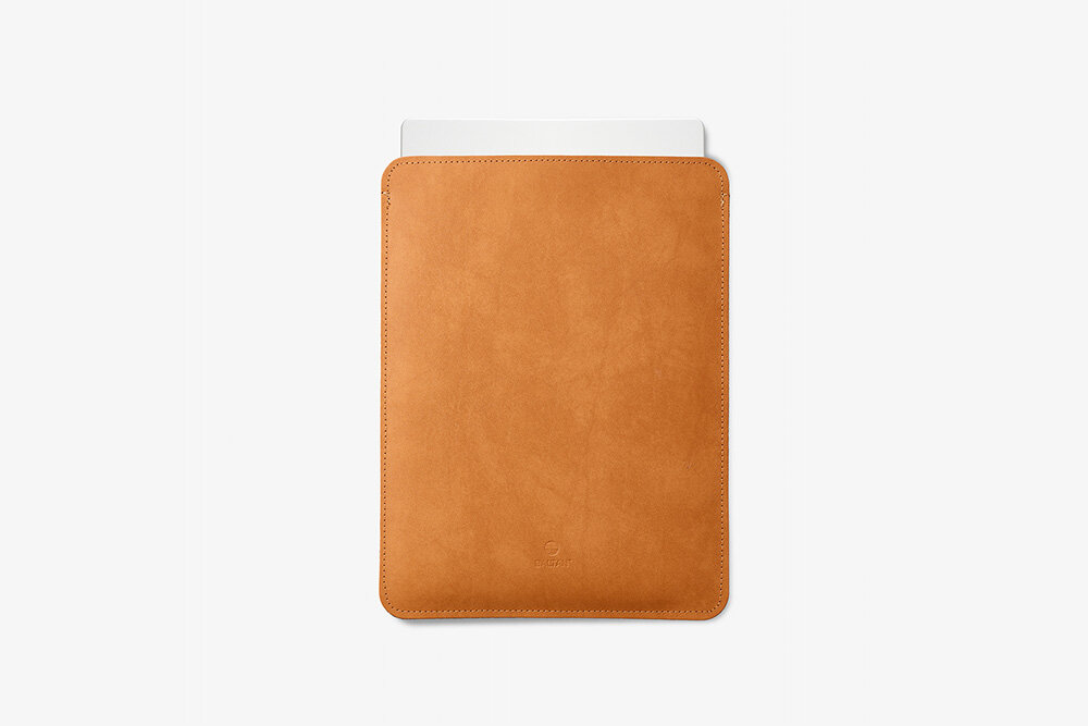 Etui na laptopa BALTAN Sleeve do Apple MacBook Air M4 15.3 cali Jasnobrązowy skóra naturalna wytrzymałość ochrona CKobieta stojąca na zewnątrz, ubrana w bluzkę w biało-czarne paski z odsłoniętymi ramionami i ciemną spódnicę, trzyma jasnobrązowe skórzane etui na laptopa. Widoczny fragment sylwetki od szyi w dół.