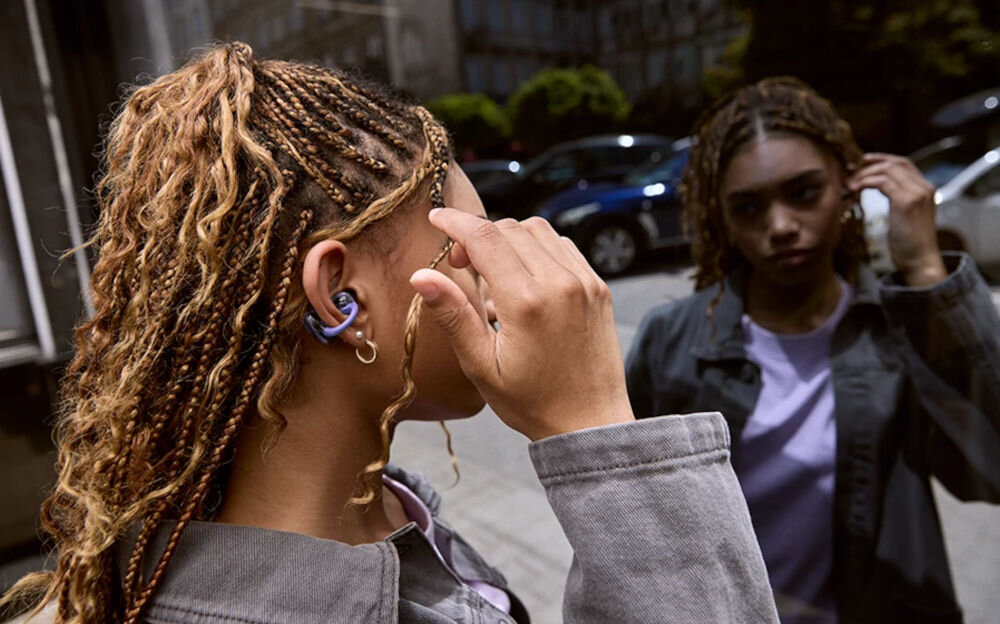 Słuchawki powietrzne JBL Soundgear Clips z pałąkiem z pamięcią kształtu i miękkim wykończeniem, osoba z założonymi słuchawkami przeglądająca się w szybie