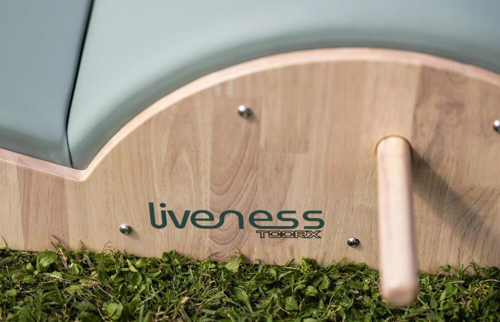 Drewniana część łuku do pilatesu z widocznym logo \'liveness TOORX\', umieszczona na trawie. Obok znajduje się drewniany uchwyt.