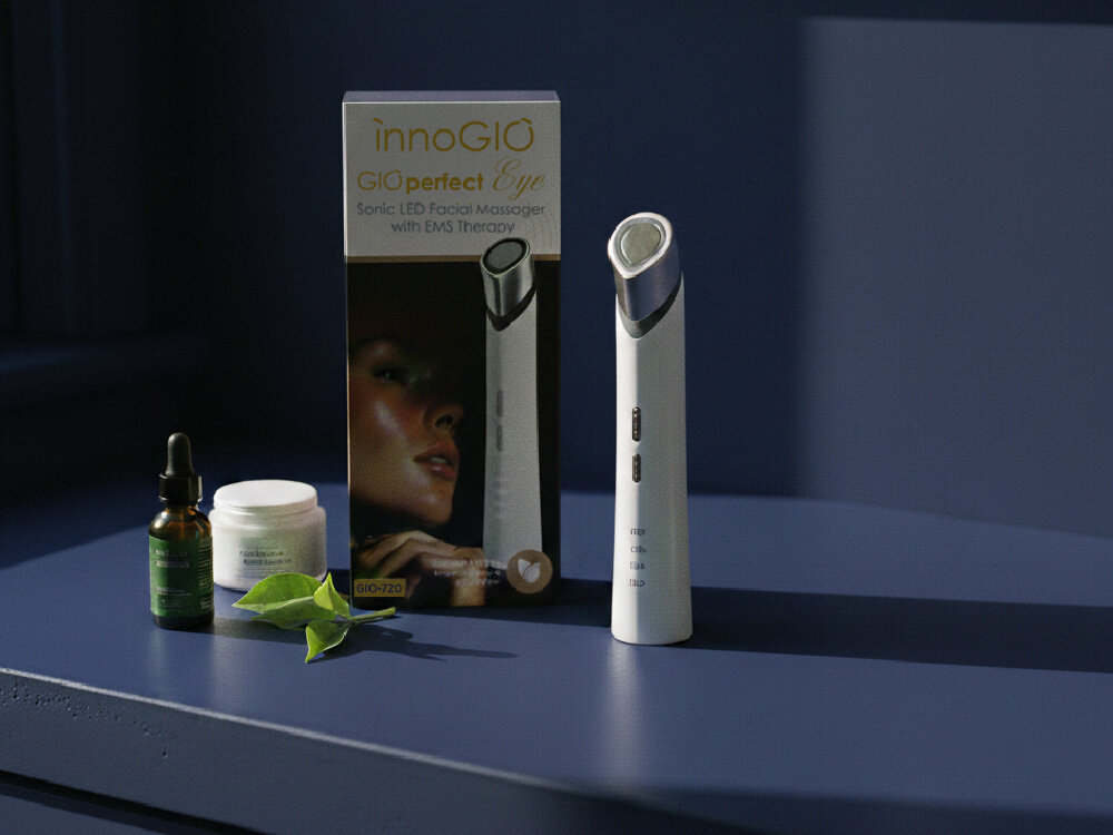 Na obrazie znajduje się masażer pod oczy oraz jego opakowanie z napisem \'innoGIO GIOperfect Eye Sonic LED Facial Massager with EMS Therapy\'. Obok masażera widoczne są buteleczka z pipetą, słoiczek oraz zielony liść.