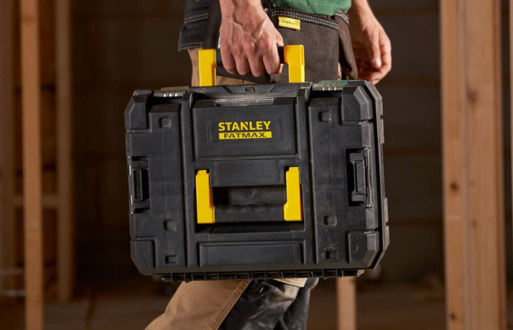Zestaw elektronarzędzi STANLEY Fatmax SFMCK211D2T-QW pomieszczenie osoba niesie walizkę w dłoni w tle drewniana ściana walizka PRO-STACK wygodne i bezpieczne przechowywanie elementy pozostają w jednym miejscu łatwo dostępne chronione przed uszkodzeniami