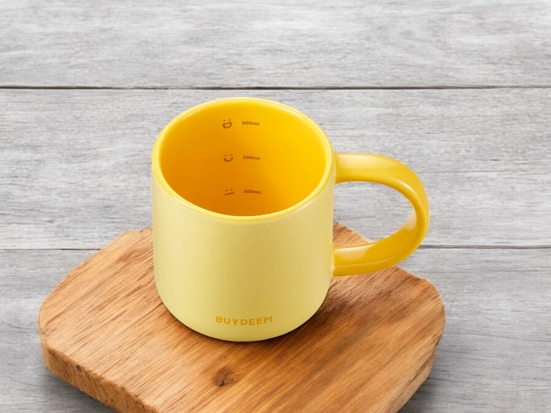 Żółty ceramiczny kubek z napisem \'BUYDEEM\' na drewnianej podstawce. Wewnątrz kubka znajdują się oznaczenia pojemności: 100ml, 200ml, 300ml.