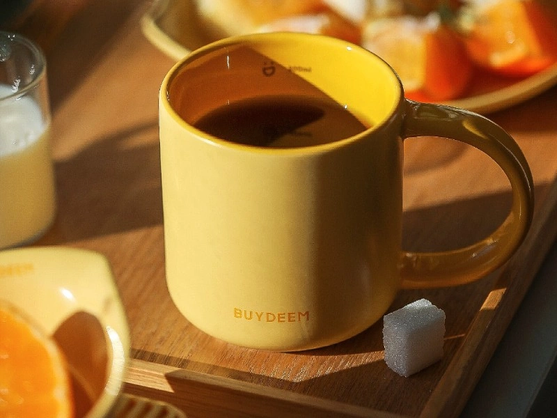 Żółty ceramiczny kubek z napisem \'BUYDEEM\' stoi na drewnianej tacy obok kostki cukru. W tle widoczne są kawałki pomarańczy i szklanka z mlekiem.