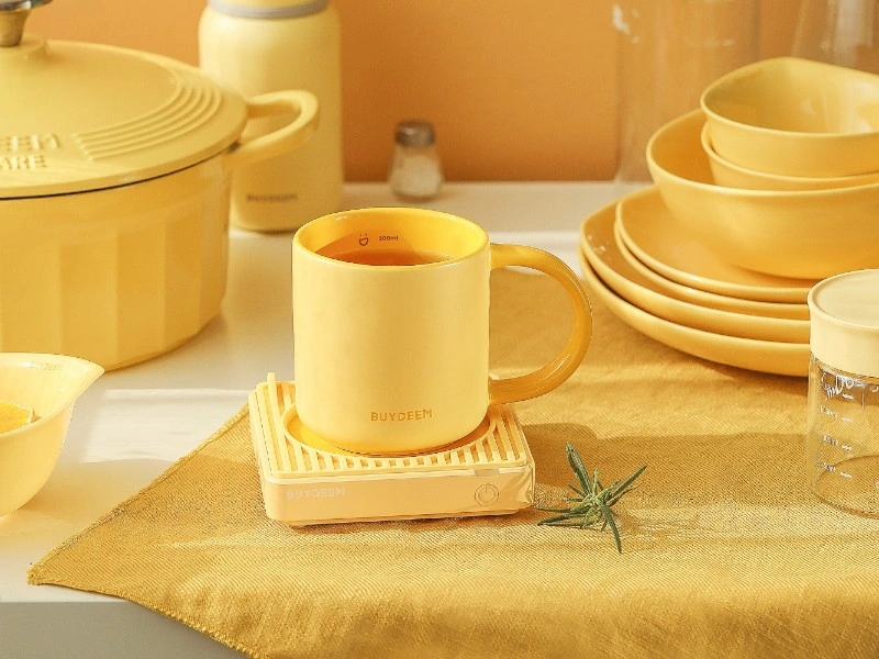 Na obrazie widoczny jest żółty kubek ceramiczny z napisem \'BUYDEEM\', stojący na podstawce. W tle znajdują się inne naczynia w podobnym kolorze oraz elementy kuchenne.