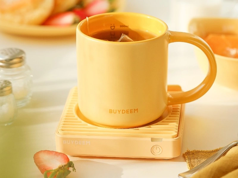 Żółty kubek ceramiczny z napisem \'BUYDEEM\' stoi na podgrzewaczu z przyciskiem zasilania. W tle widoczne są elementy zastawy stołowej i plasterek truskawki.